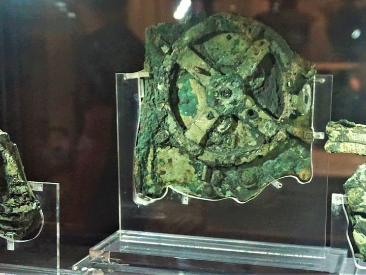 Green corroded bronze fragments of the Antikythera mechanism mounted on clear stands in a glass case at the National Archaeological Museum in Athens