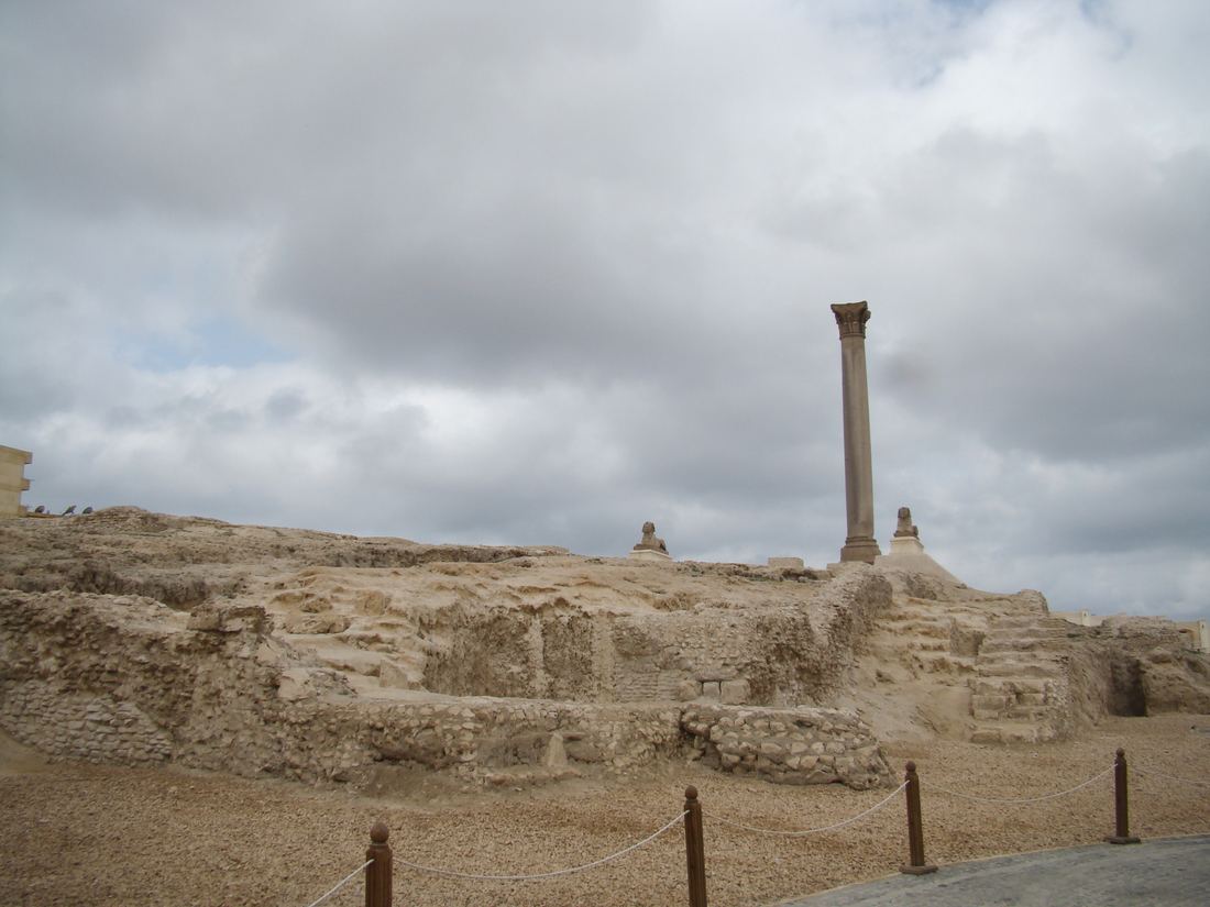 The tall granite column known as Pompey's Pillar rising above the excavated stone ruins of the Serapeum in Alexandria under a cloudy sky