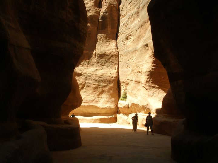 The narrow Siq canyon at Petra, Jordan, with towering curved walls of pink and orange sandstone and two small figures walking toward a band of light